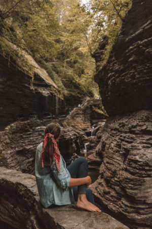 A woman in a blue shirt sitting in a dark rocky ravine trees lining the top of the ravine at rainbow falls, Finger Lakes New York