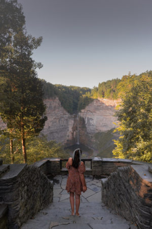 A woman in a dress standing at a stone viewing platform with a large rocky waterfall in the distance surrounded by trees